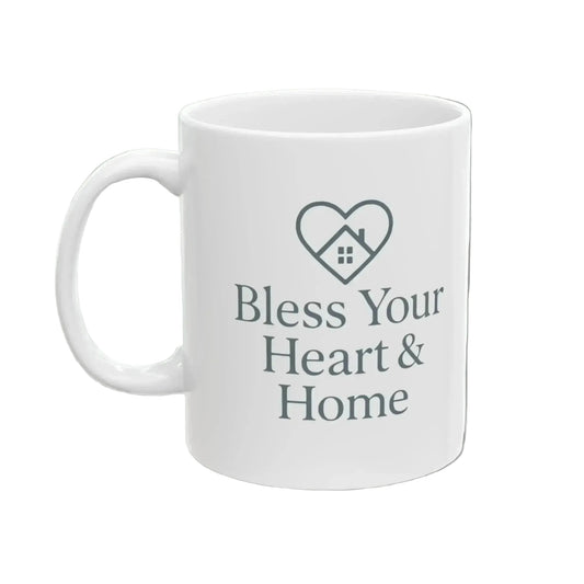 A white ceramic mug with a gray printed design featuring a heart outline with a small house inside and the text 'Bless Your Heart & Home' on the front.