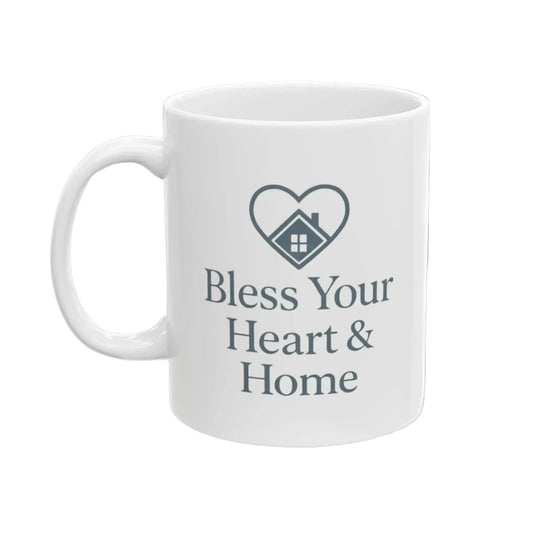 A white ceramic mug with a dark gray printed design featuring a heart outline enclosing a small house and the text 'Bless Your Heart & Home'.