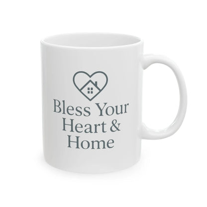 A white ceramic mug with a dark gray printed design featuring a heart outline containing a small house and the text 'Bless Your Heart & Home'.