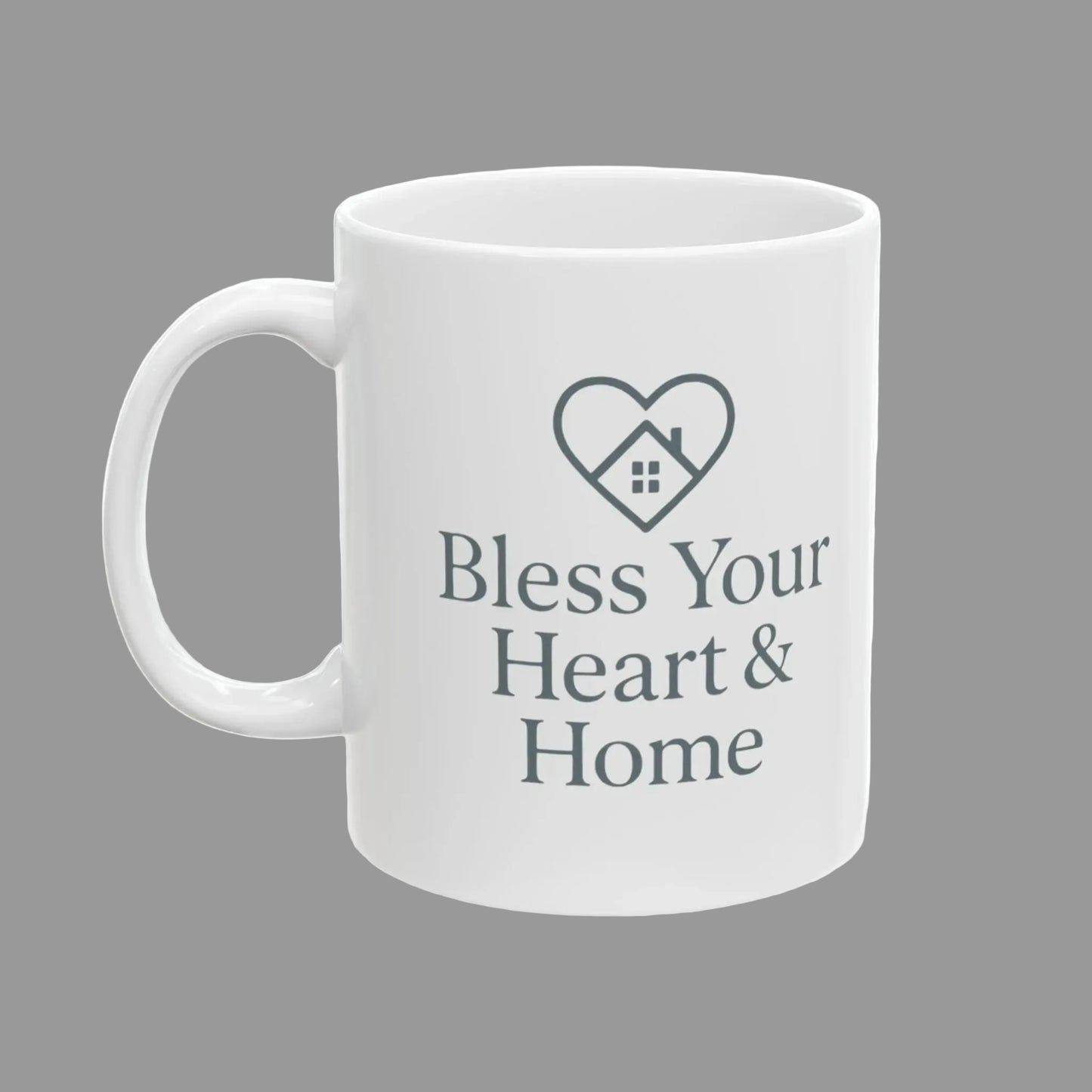 A white ceramic mug with a gray printed design featuring a heart outline with a small house inside and the text 'Bless Your Heart & Home' on the front.