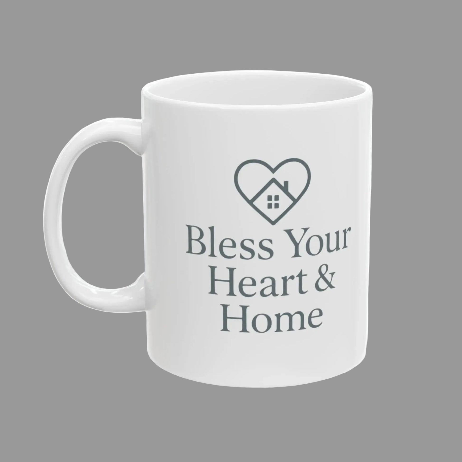 A white ceramic mug with a gray printed design featuring a heart outline with a small house inside and the text 'Bless Your Heart & Home' on the front.