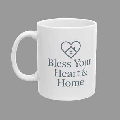 A white ceramic mug with a gray printed design featuring a heart outline with a small house inside and the text 'Bless Your Heart & Home' on the front.