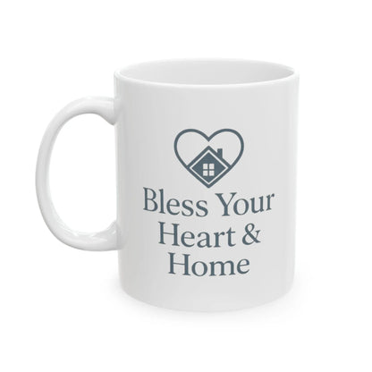 A white ceramic mug with a dark gray printed design featuring a heart outline enclosing a small house and the text 'Bless Your Heart & Home'.