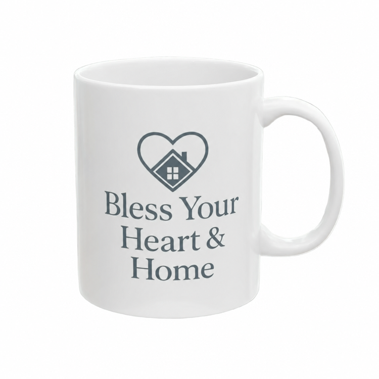 A white ceramic mug with a dark gray printed graphic of a heart containing a house, above the text 'Bless Your Heart & Home'.