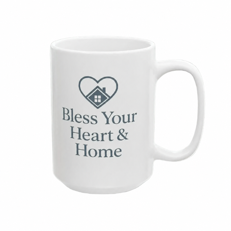 A white ceramic mug with a dark gray printed design featuring a heart outline enclosing a small house and the text 'Bless Your Heart & Home'.