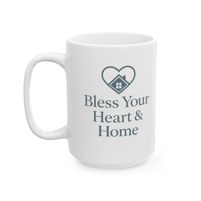 A white ceramic coffee mug with a dark gray printed design featuring a heart-shaped house icon and the text 'Bless Your Heart & Home'.