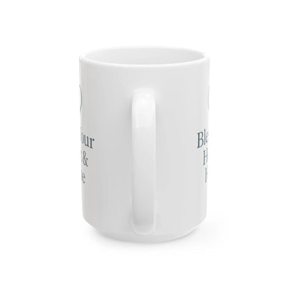 A white ceramic mug with a handle, shown from a side angle on a white background. The mug features dark gray text partially visible on the sides, including the words 'Bless' and 'H.