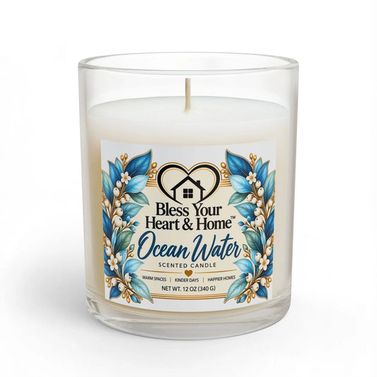 A 11 oz scented candle in a clear glass jar with a white wax fill and a central wick. The front label features blue and gold floral illustrations with the text 'Bless Your Heart & Home Ocean Water SCENTED CANDLE'.