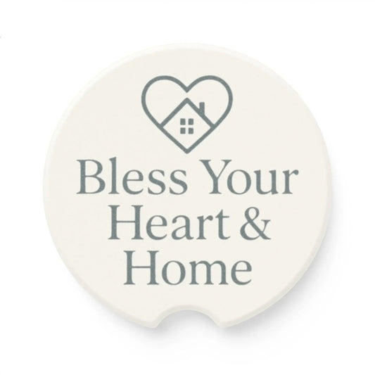 A round white coaster with a small cutout at the bottom, featuring a gray heart outline containing a house icon and the text 'Bless Your Heart & Home' in gray. A small 'Comfort Colors' logo is visible in the top-left corner.