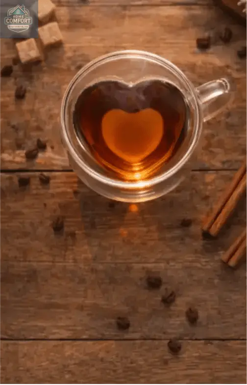 Heart-shaped glass mug filled with tea on a wooden surface with cinnamon sticks and sugar cubes.