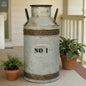 Vintage-style metal milk can with 'No 1' label on a concrete surface, surrounded by plants.