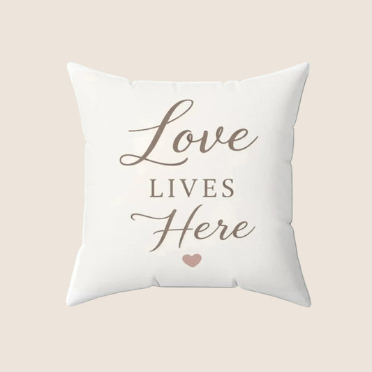 A square white throw pillow featuring the phrase 'Love LIVES Here' in brown script and serif lettering, with a small pink heart underneath, shown on a clean white background.